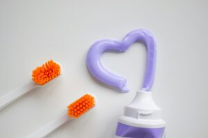 Photo by Ann Zzz: https://www.pexels.com/photo/a-toothbrushes-and-toothpaste-on-a-white-surface-8191884/ Orange toothbrushes next to purple toothpaste in the shape of a heart